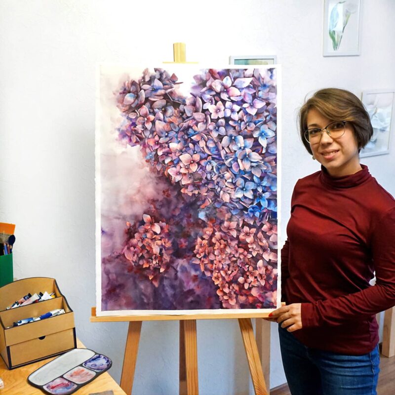 A photo of an artist next to a large watercolor painting of hortensia flowers