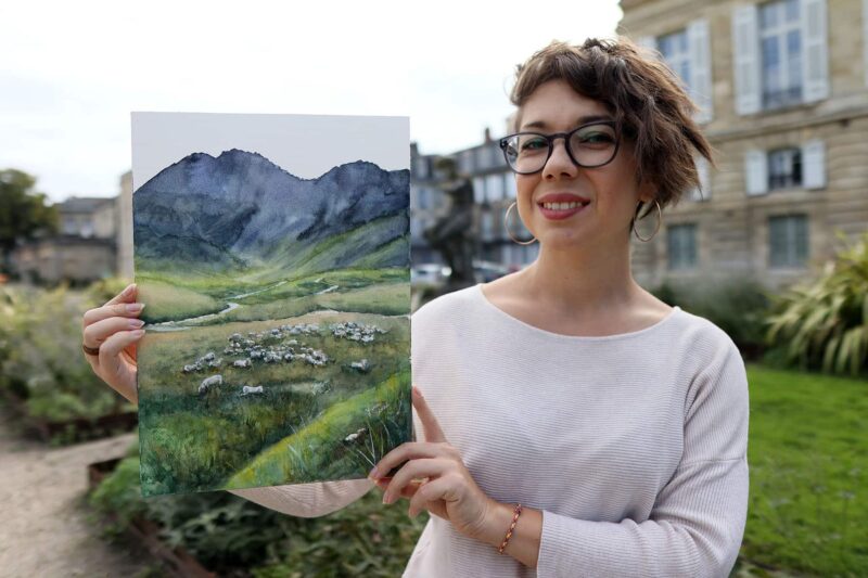 an artist holding a watercolor painting of a mountain landscape with a flock of sheep and a shepherd dog