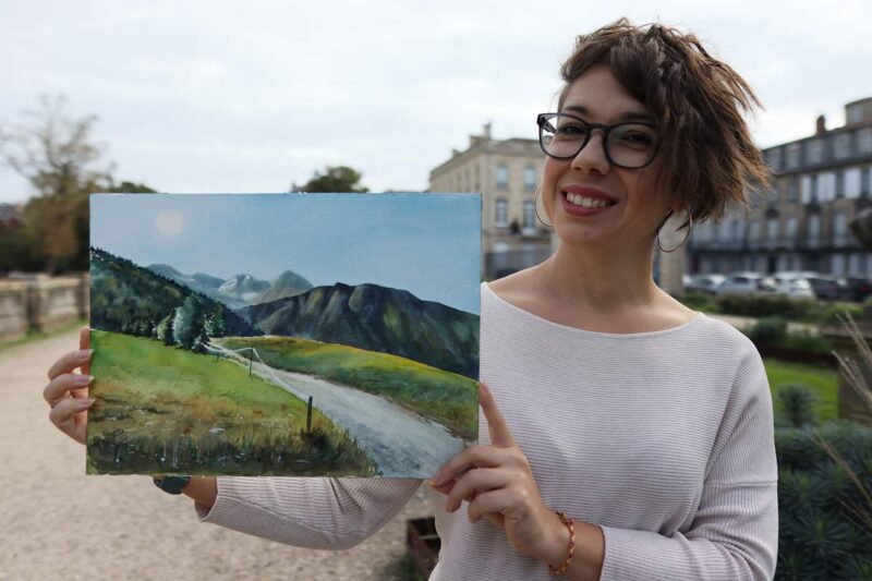 an artist holding a watercolor painting of a sunny landscape of the alps